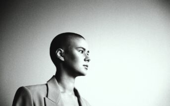 Black and white photo of a person with a shaved head, wearing a blazer. They are gazing to the right against a plain backdrop, creating a serene and contemplative mood.