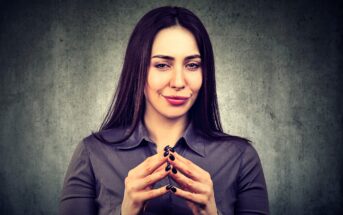 Woman with long dark hair in a gray blouse, smirking and clasping hands together with fingertips touching, stands against a textured gray background.