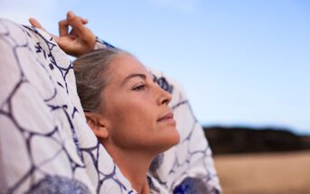 A person with grey hair is shown in close-up with their arms raised behind their head. They wear a white garment with blue patterns and appear relaxed, set against a clear blue sky and sandy beach backdrop.
