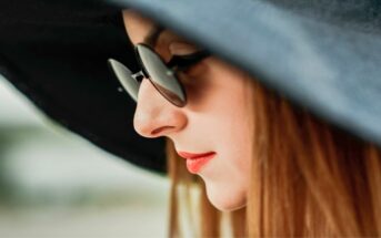 A close-up side profile of a person with long hair, wearing a large dark sun hat and round sunglasses. The individual has vibrant red lipstick and appears to be looking downward, with a blurred background.