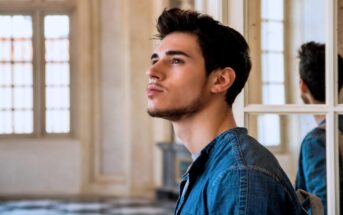 A young man with short brown hair and a slight beard is gazing upward thoughtfully. He is dressed in a blue denim shirt and stands indoors near a window with a soft light illuminating the background.