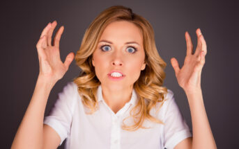A woman with blonde hair, wearing a white shirt, has an angry expression. Her eyes are wide open, mouth slightly open showing teeth, and her hands are raised with fingers spread apart. The background is dark, making her face and gestures more prominent.