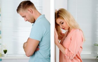 A man and a woman stand separated by a wall, both with somber expressions. The man on the left leans his head forward, arms crossed, while the woman on the right rests her head and hands against the wall. Both appear to be deep in thought or sad.