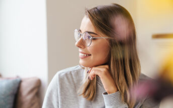 A woman with shoulder-length blond hair and glasses is smiling while looking to the side. She has her hand resting under her chin. She is indoors, with a blurred background that includes soft tones of yellow and pink.