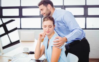 A man in a blue shirt places his hand on the shoulder of a seated woman, who looks uncomfortable and is pulling away. They are in an office setting with a computer and papers on the desk. The interaction appears tense.