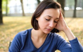 A young woman with short brown hair, wearing a blue long-sleeve shirt, rests her head on her hand while sitting outdoors in a park. She appears to be deep in thought, with her eyes closed and a neutral expression. The background is blurred with trees and fallen leaves.