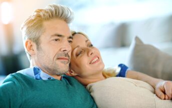 A middle-aged couple is sitting closely together on a couch, appearing content and relaxed. The man, with gray hair and a beard, is wearing a teal sweater over a blue shirt. The blonde woman is leaning on his shoulder, smiling gently in a light-colored top.