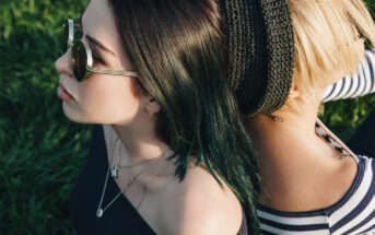 Two people sit back-to-back on grass. One has dark hair and wears round sunglasses and an off-the-shoulder top. The other has blond hair under a black knitted hat and wears a striped top. Both have necklaces and appear relaxed with eyes closed.