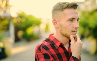 A young man with blonde hair and a beard, wearing a red and black checkered shirt, stands outdoors on a sunny day. He is looking to the side with one hand thoughtfully touching his chin. The background is out of focus, featuring greenery and a blurred pathway.