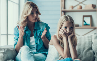 A woman and a young girl sit on a couch in a living room. The woman, with blonde hair, is speaking and gesturing with her hands, while the girl, also blonde, rests her head on her hands, looking upset. Shelves with decor items are in the background.