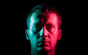 A man is photographed against a black background with dramatic lighting. The left side of his face is illuminated with blue light, while the right side is illuminated with red light. The contrast creates a striking visual effect. He has a serious expression.