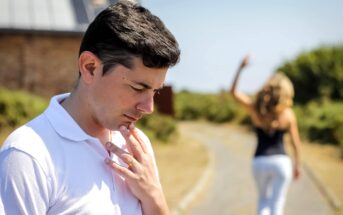 A man in a white polo shirt stands in the foreground with a thoughtful or upset expression, touching his chin. In the blurred background, a woman with long blonde hair walks away on a path. Trees and a building are visible in the distance.