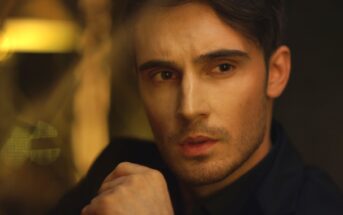 A man with short brown hair and a trimmed beard looks contemplatively to his right. He wears a dark jacket, and his hand is placed near his chin. The background is softly lit with warm tones, giving the image a moody and reflective atmosphere.