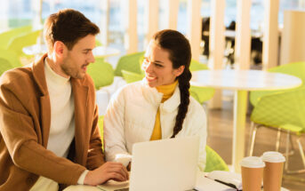 A man and a woman sit together at a table with a laptop in front of them. The man wears a brown coat and the woman has a white jacket, and they are smiling at each other. Two coffee cups are on the table, and the setting appears to be a bright, modern cafe.