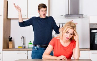 A man with a frustrated expression is standing in a kitchen, gesturing with his hands. A woman with a downcast expression sits at a table in the foreground, leaning her head on one hand. The scene suggests they are having a disagreement.