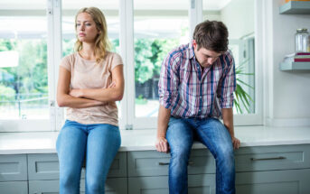 A woman and a man sitting on a kitchen counter, both facing forward. The woman has her arms crossed and looks away with a stern expression. The man has his hands on his lap, leaning forward slightly, looking down. The atmosphere appears tense.