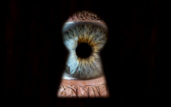 A close-up of a human eye seen through a keyhole. The eye has a bright blue and yellow iris with a dark black pupil in the center, surrounded by the dark edges of the keyhole. The surrounding area is mostly black, focusing the viewer's attention on the eye.