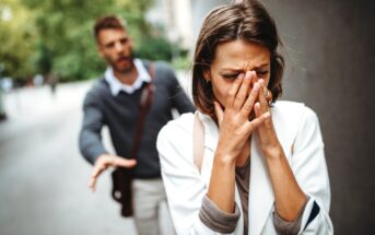 A woman in distress covers her face with her hands, walking away from a man who appears to be trying to talk to her. The man has an outstretched arm and a concerned expression. They are outdoors, with blurred greenery in the background.