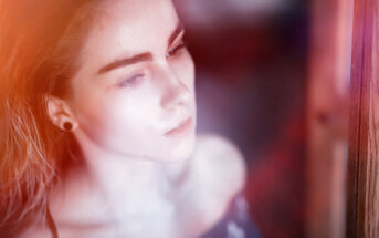 A close-up of a person with a contemplative expression, looking out a window with soft, natural light illuminating their face. The image has a dreamy, ethereal quality with a slight blur and warm tones. The person has long hair and is wearing a dark, off-the-shoulder top.