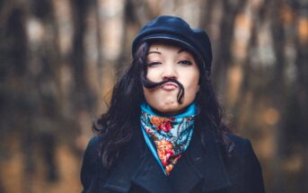 A person with dark hair is wearing a black cap and a colorful scarf. They have playfully positioned their hair over their upper lip to resemble a mustache. The background is blurred with dark, earthy tones.