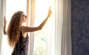 A woman with long curly hair and wearing glasses pulls back white curtains to let sunlight into a room with dark textured walls. She is standing beside a large window, dressed in a colorful sleeveless top. The scene exudes a calm and peaceful atmosphere.