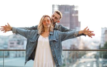 A couple stands on a rooftop with the city skyline in the background. The woman in a white dress and denim jacket spreads her arms out, while the man behind her, wearing a denim jacket, gently holds her arms. Both are smiling and enjoying the view.