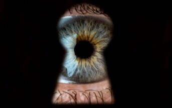 Close-up of a human eye with a greenish-brown iris, seen through the keyhole of a door. The surrounding area appears dark, focusing attention on the detailed texture and color variations in the eye.