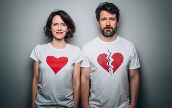 A woman and a man stand side by side against a gray wall. The woman is smiling and wears a shirt with a red heart, while the man looks serious and wears a shirt with a red broken heart.