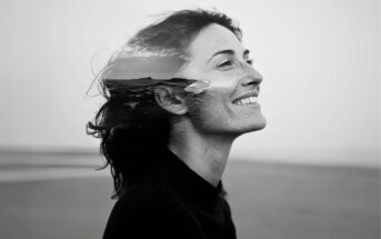 A black and white double exposure photo of a woman smiling in profile, with her hair blending into an ocean scene featuring waves and clouds.