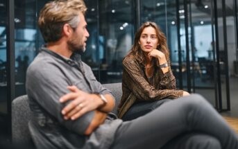 A man and a woman sit on a sofa in a modern office, engaged in a serious conversation. The woman looks attentively at the man, who has his arms crossed. Glass walls and office furniture are visible in the background.
