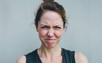 A woman with light skin and brown hair in a bun is wearing a black sleeveless shirt and scrunching her face, expressing displeasure or annoyance, against a plain light gray background.