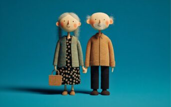 Two handcrafted dolls of an elderly woman and man stand side by side against a blue background. The woman holds a basket and wears a dress and cardigan; the man wears a brown jacket and dark pants. Both are smiling.
