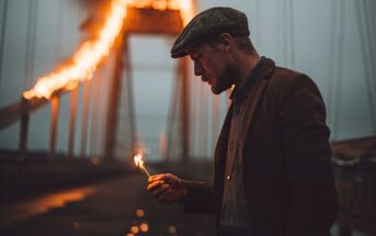 A man in a flat cap and jacket holds a lit match on a bridge at dusk, while large flames burn dramatically along the bridge’s cables in the background.