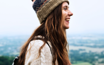 A woman with long hair is smiling, wearing a knit beanie and sweater, standing outdoors. The background is a blurred view of a landscape with greenery and sky.