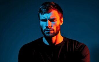 A man with a beard and mustache looks intensely at the camera, lit dramatically with blue and orange lighting on his face, wearing a black shirt against a dark background.