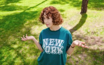A person with curly hair is standing on a grassy area, wearing a green shirt with "New York" printed on it. They have an expression of uncertainty, with their hands raised and shoulders slightly shrugged. Shadows of trees are visible on the grass.