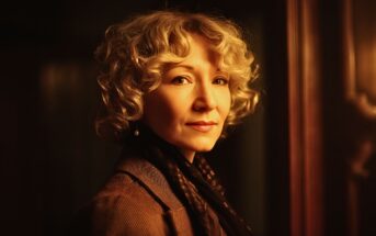 A woman with short curly blonde hair and light skin stands indoors, gazing gently at the camera. She wears a brown jacket and scarf, with warm lighting highlighting her face against a dark background.