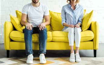 A man and woman sit on a bright yellow couch, both wearing white sneakers. The man wears a white T-shirt and jeans, while the woman wears a blue-striped shirt and white pants. They look straight ahead, not touching, against a white brick wall.