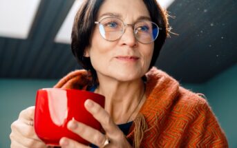 A person with short dark hair and glasses is holding a red mug while wearing an orange patterned shawl. They are looking thoughtfully to the side, with light coming from a skylight above.