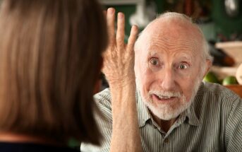 An elderly man with white hair and a beard gestures expressively with his hand while talking to a woman whose back is to the camera. The man appears engaged and animated in conversation.