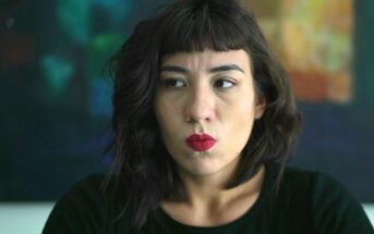 A woman with dark wavy hair and red lipstick looks to the side with a serious expression. She is wearing a black top, and the background is blurred with abstract colors.