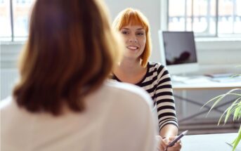A woman with short red hair and a striped shirt smiles while holding a pen, sitting across a desk from another woman in an office with large windows and a computer in the background.