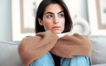 A woman with long dark hair sits on a couch, resting her chin on her crossed arms. She wears a brown sweater and blue jeans, and looks thoughtfully to the side. The background is softly blurred.