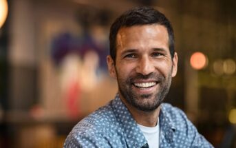A man with short dark hair and a beard smiles warmly at the camera, wearing a blue patterned shirt. The background is softly blurred, suggesting an indoor setting with ambient lighting.