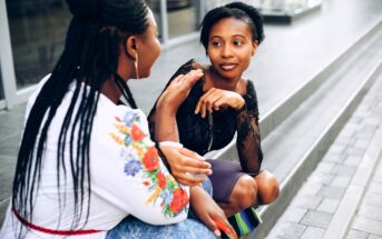 Two women sit on outdoor steps, engaged in a lively conversation. One woman gestures with her hand while the other listens attentively, smiling. Both are casually dressed, and the scene is bright and relaxed.