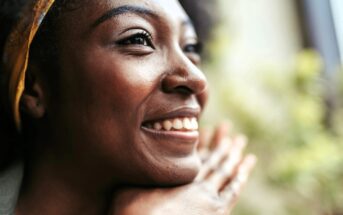 A close-up of a smiling woman resting her chin on her hands, looking off to the side with a joyful expression. Bright natural light highlights her face, and a blurred, green background is visible.