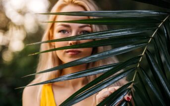 A woman with long blonde hair stands behind green palm leaves, with her face partially obscured. She is wearing a yellow top and has red painted nails. The background is softly blurred with natural light.