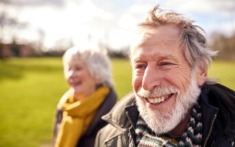 An older man with a gray beard smiles outdoors, wearing a striped scarf and jacket. A woman with gray hair and a yellow scarf stands slightly behind him. Both appear happy in a sunlit, grassy park.