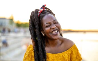 A woman with long, natural hair and a red headband smiles brightly while standing outdoors near water at sunset, wearing an off-the-shoulder yellow top.