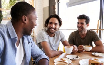 Three men sit at a café table, smiling and talking while holding coffee cups. Sunlight streams in through large windows, creating a warm and cheerful atmosphere.